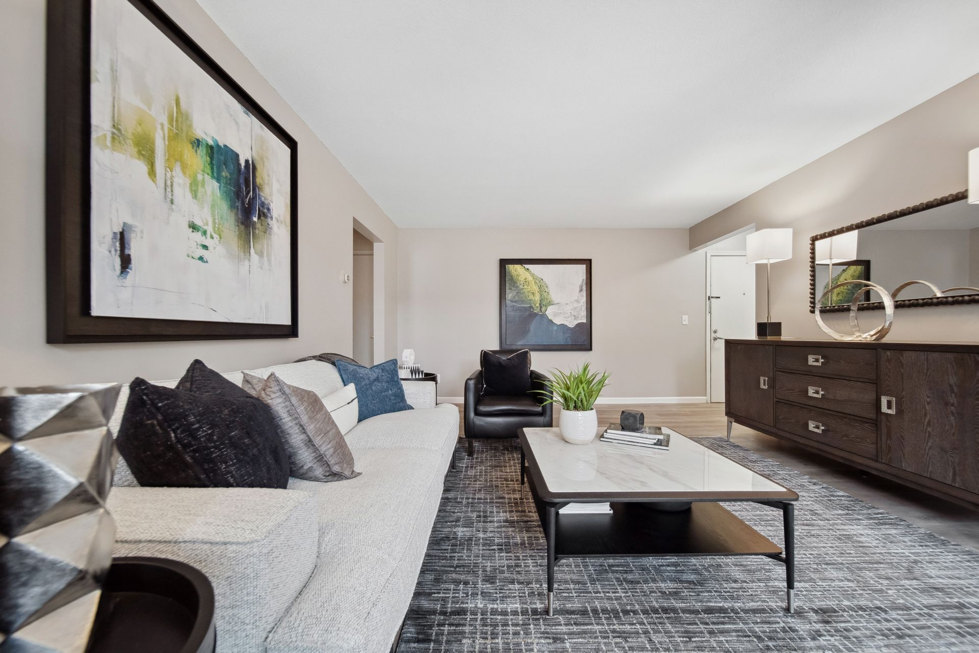 Bright modern living room with white sofa, black chair, gray rug, and dark wood dresser open to kitchen area