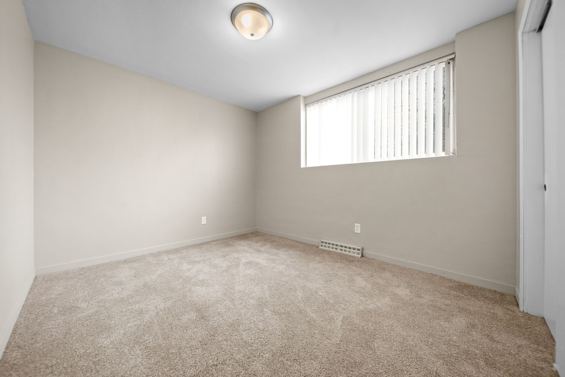 Empty beige carpeted bedroom with white walls, ceiling light, and a small window with vertical blinds