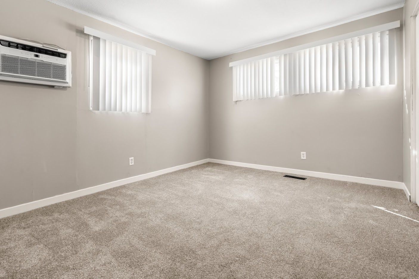 Empty beige room with carpet, two windows with blinds, and a wall air conditioner