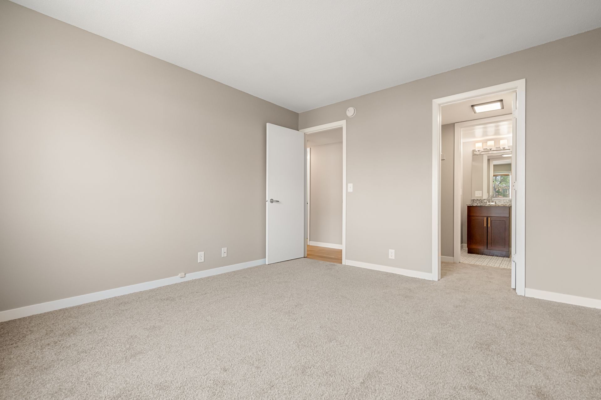 Empty beige bedroom with carpet, white doors, and an attached bathroom visible through the open doorway.