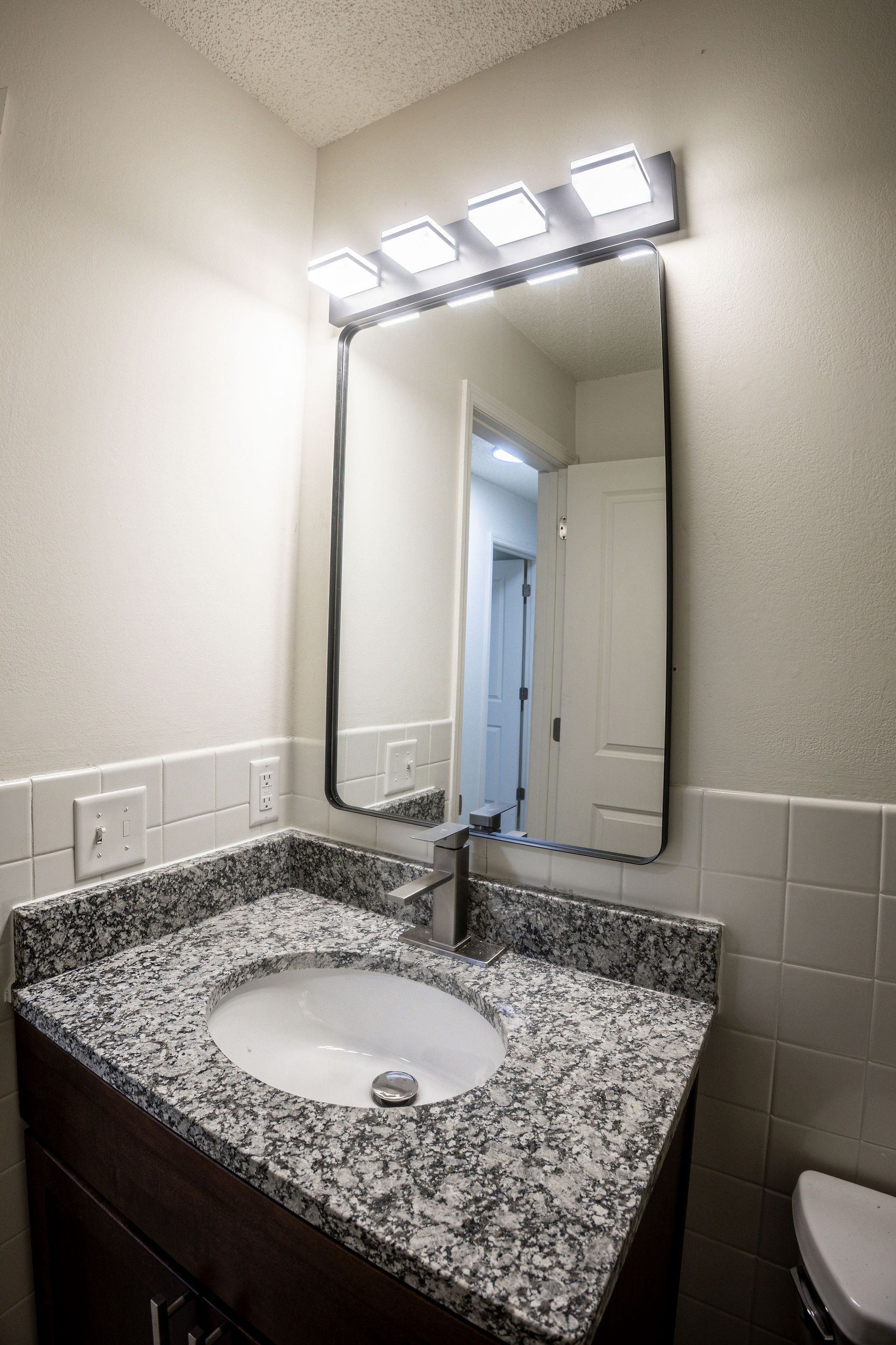 Granite bathroom vanity with sink and mirror under bright lights, showing a doorway reflected in the mirror