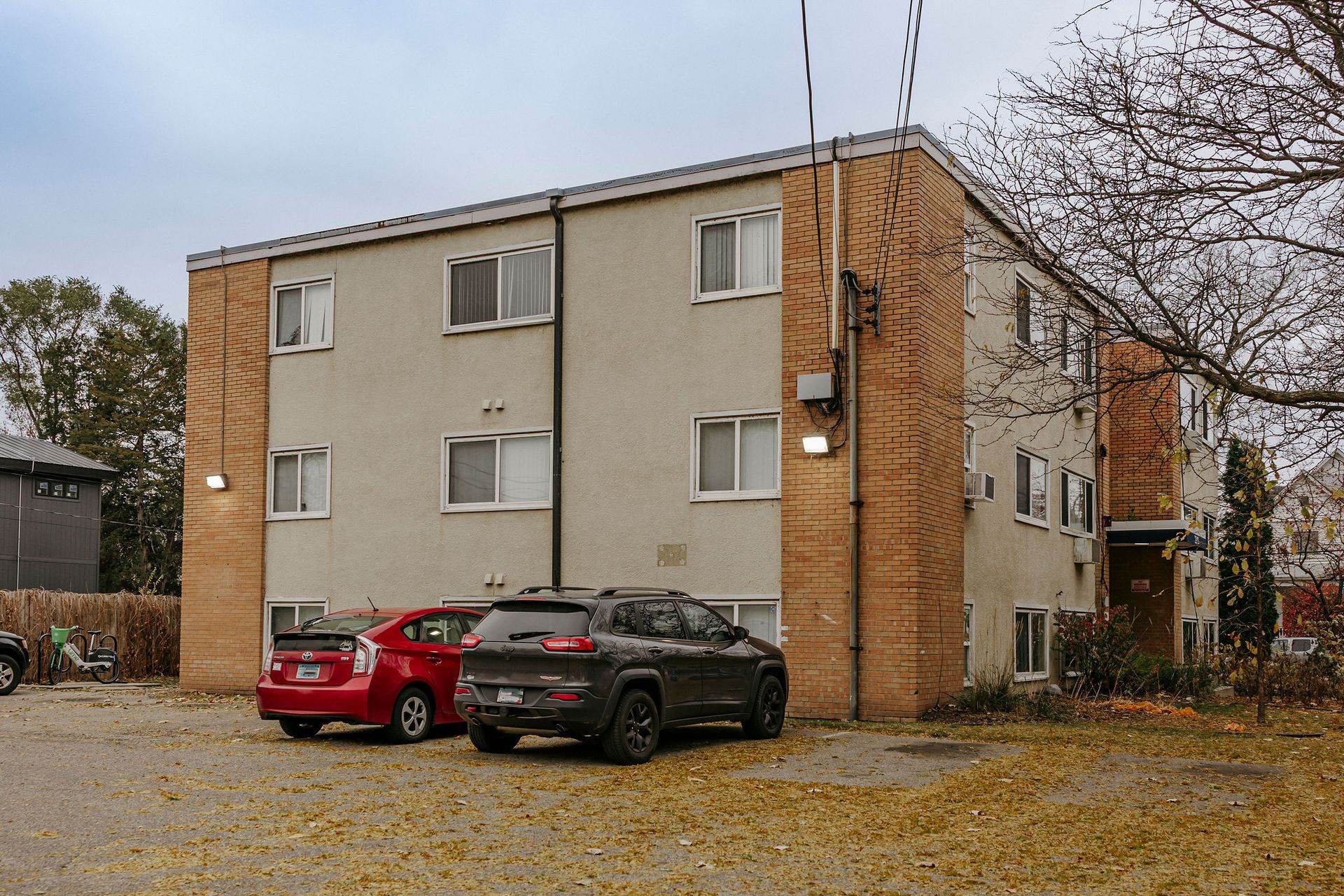 Apartment building exterior with two parked cars on a gravel lot under overcast skies