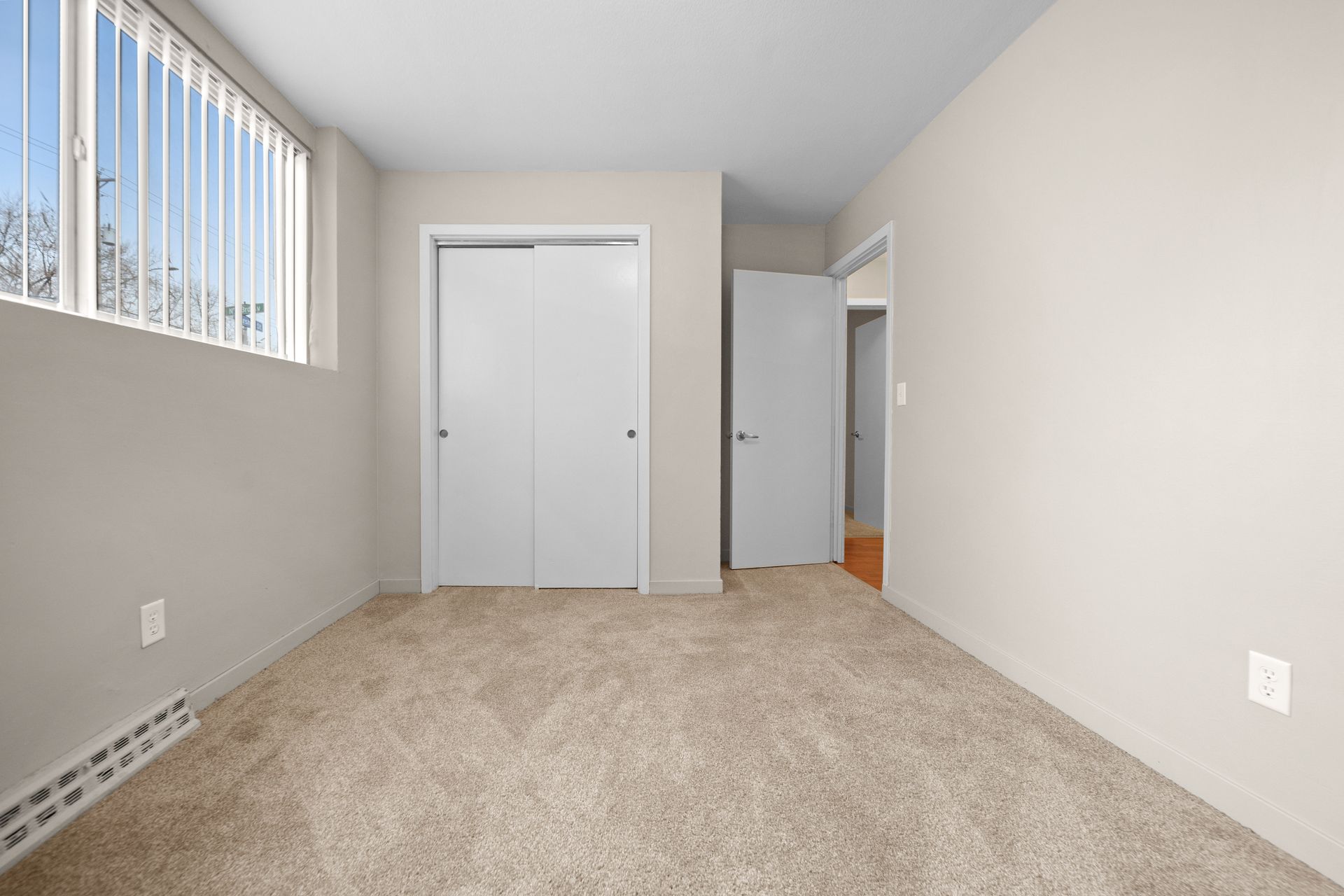 Empty beige bedroom with carpet, closet doors, window blinds, and an open doorway