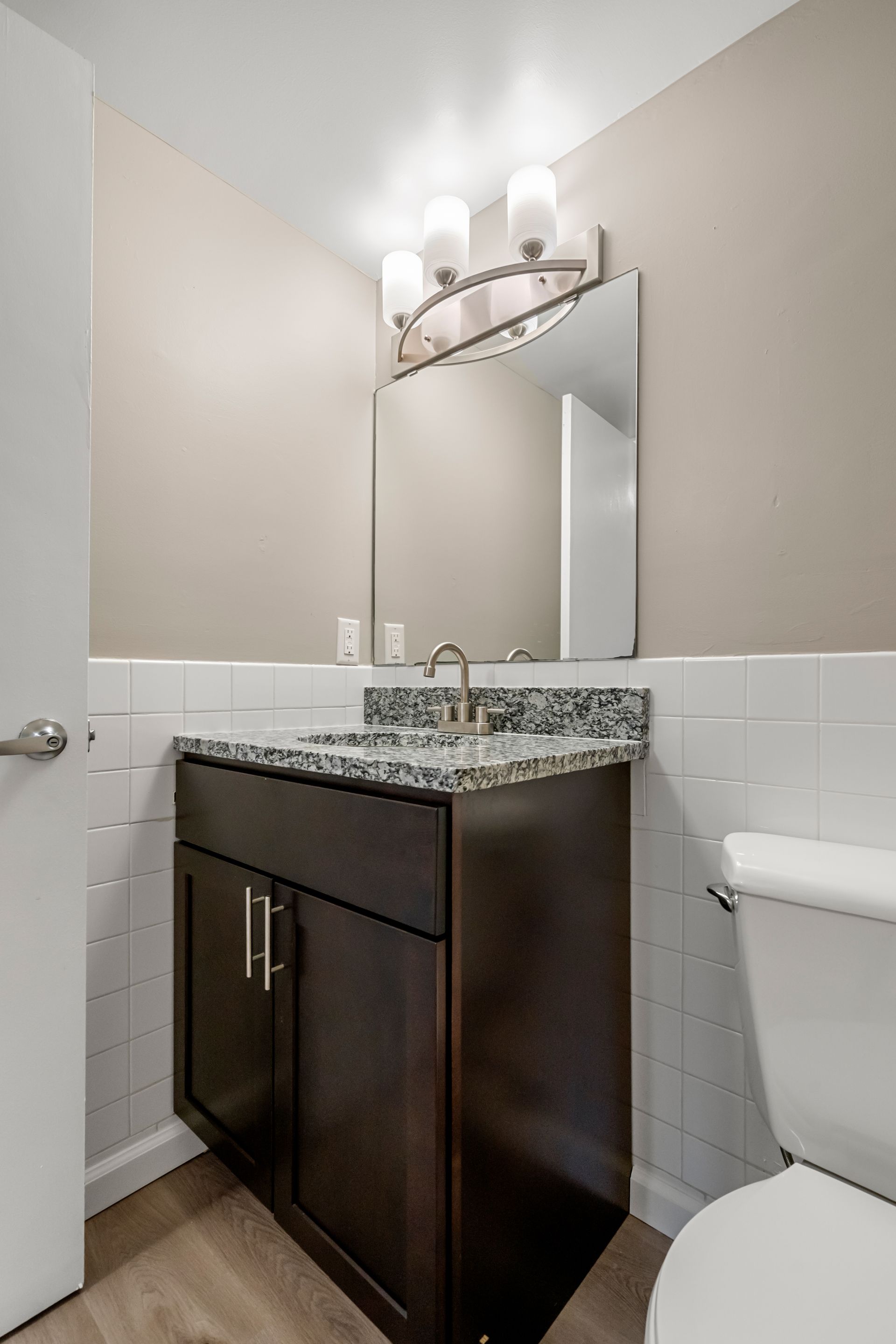 Small bathroom with dark wood vanity, speckled countertop, mirror, sconce lights, and part of a toilet.