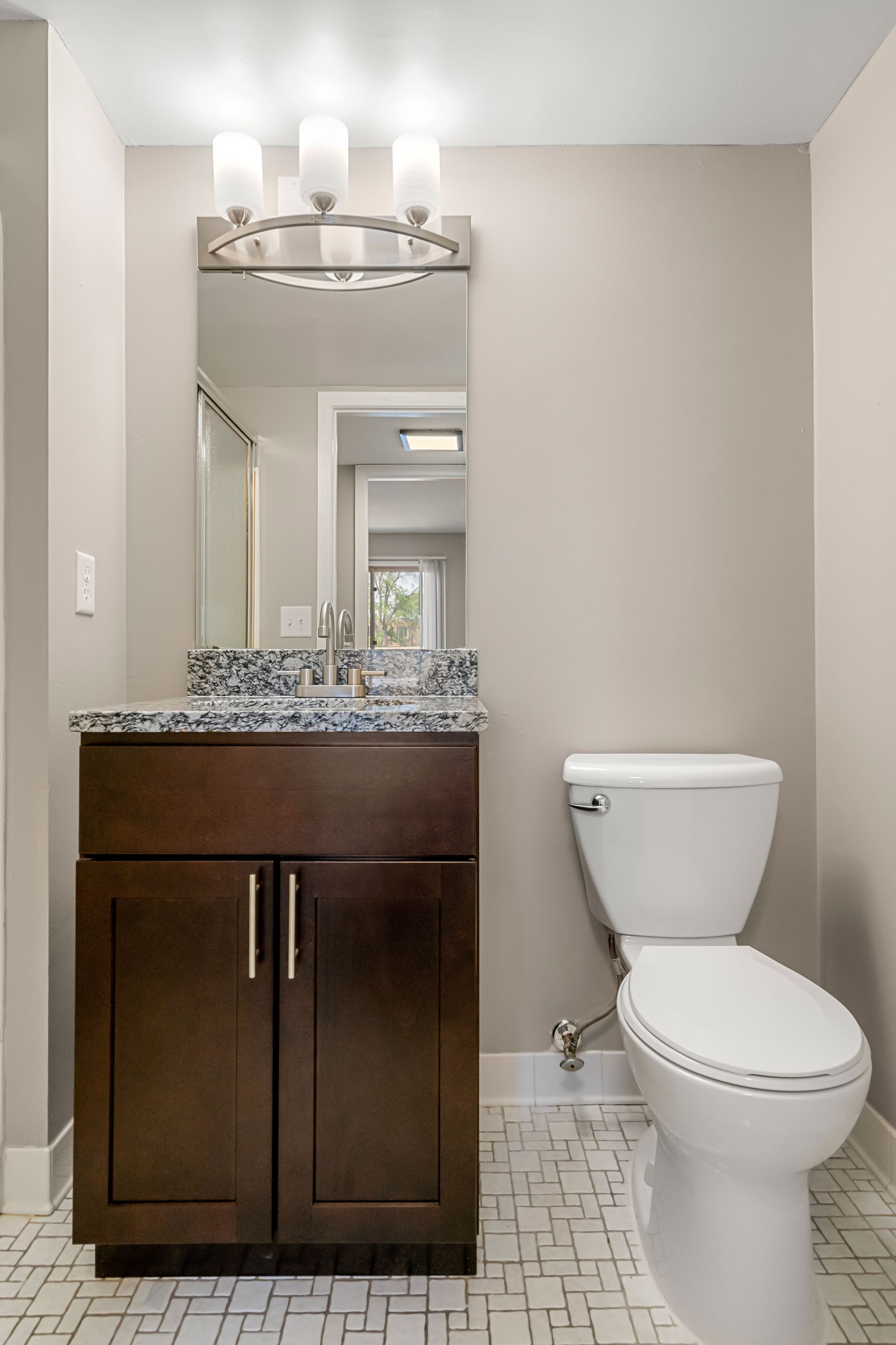Small bathroom with dark wood vanity, mirror, light fixture, and white toilet on patterned tile floor.