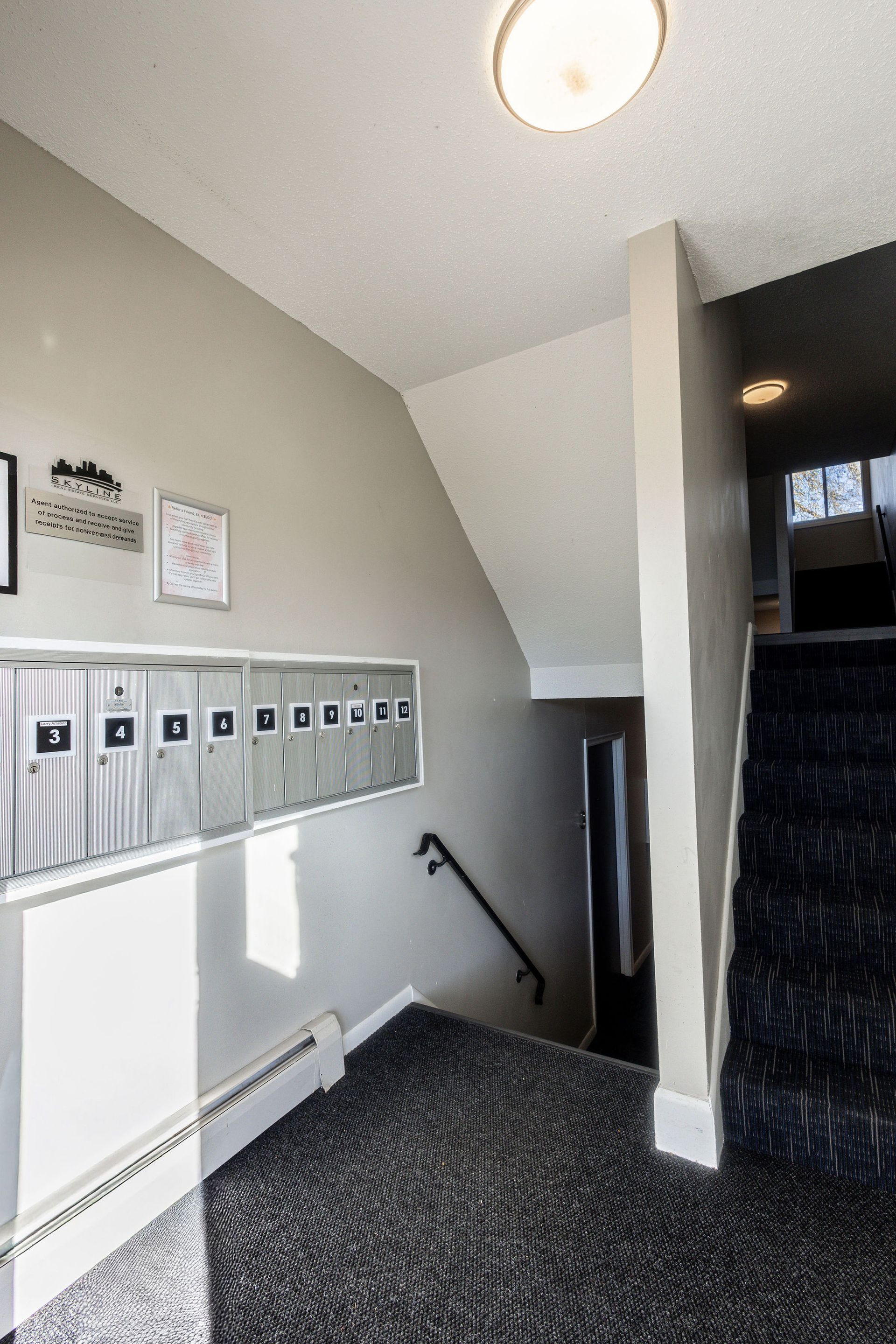 Apartment hallway with mailboxes, stairs, and wall art, lit by a ceiling light