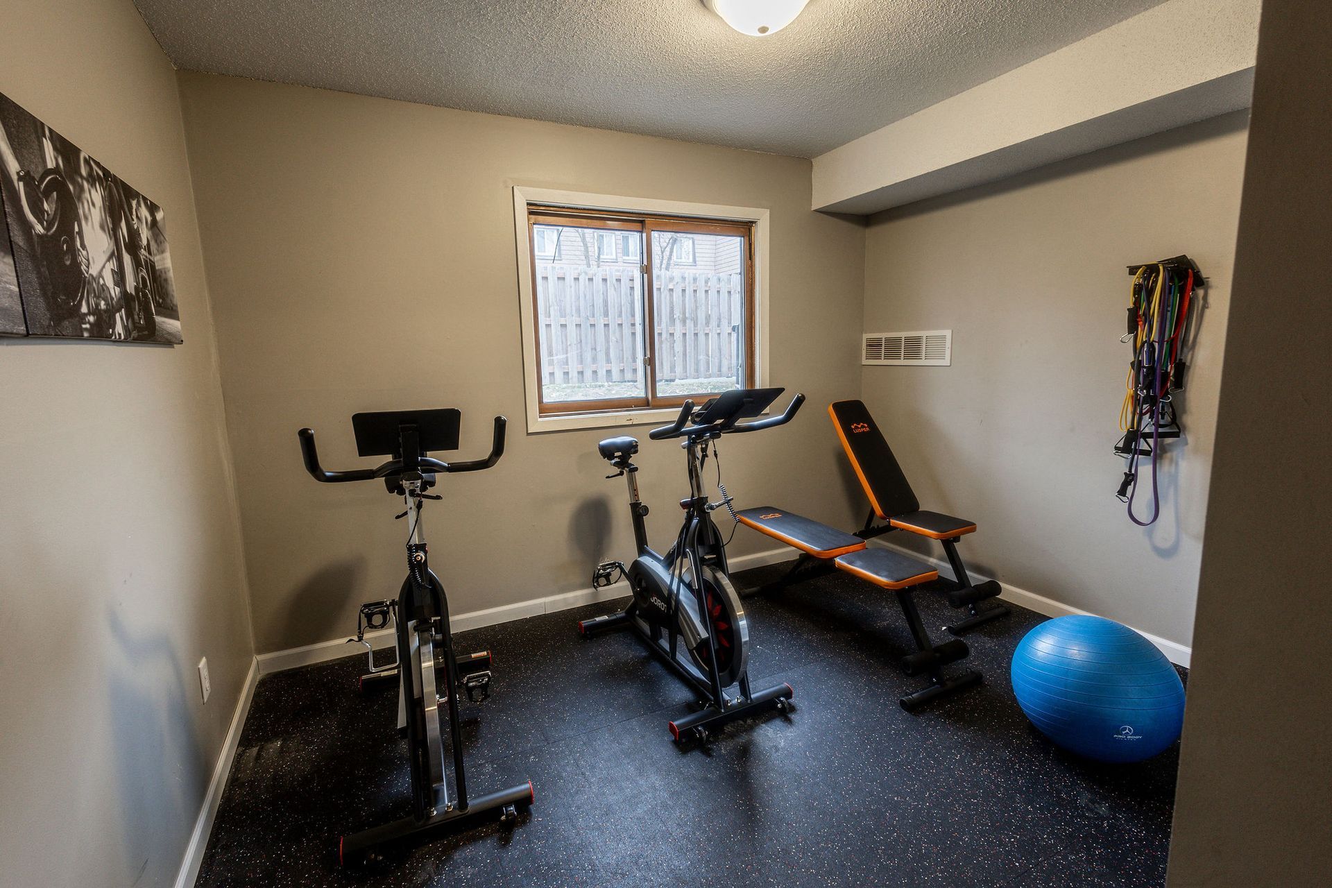Small home gym with exercise bike, rowing machine, weights, and blue exercise ball in a carpeted room.