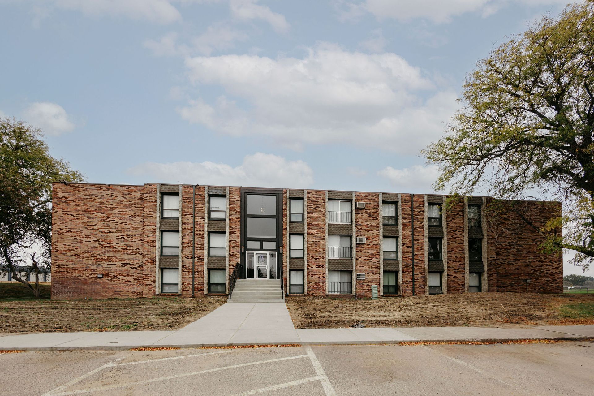 Brick apartment building with central entrance, flanked by trees and a parking lot in front