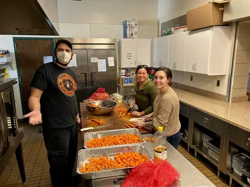 Volunteers in a kitchen stand beside trays of prepared food and bowls on a long counter.