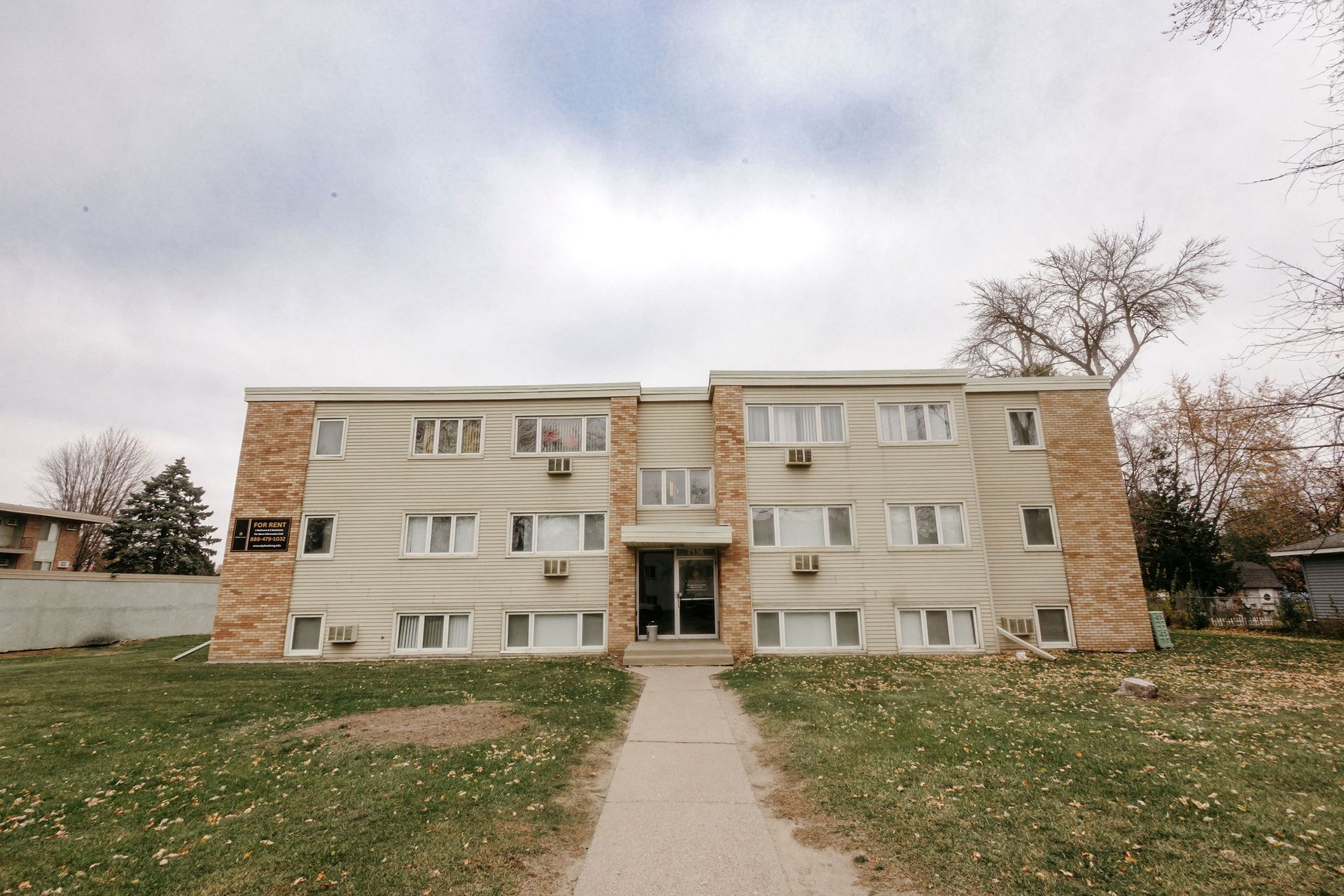 Three-story apartment building with tan siding and a central walkway under a cloudy sky