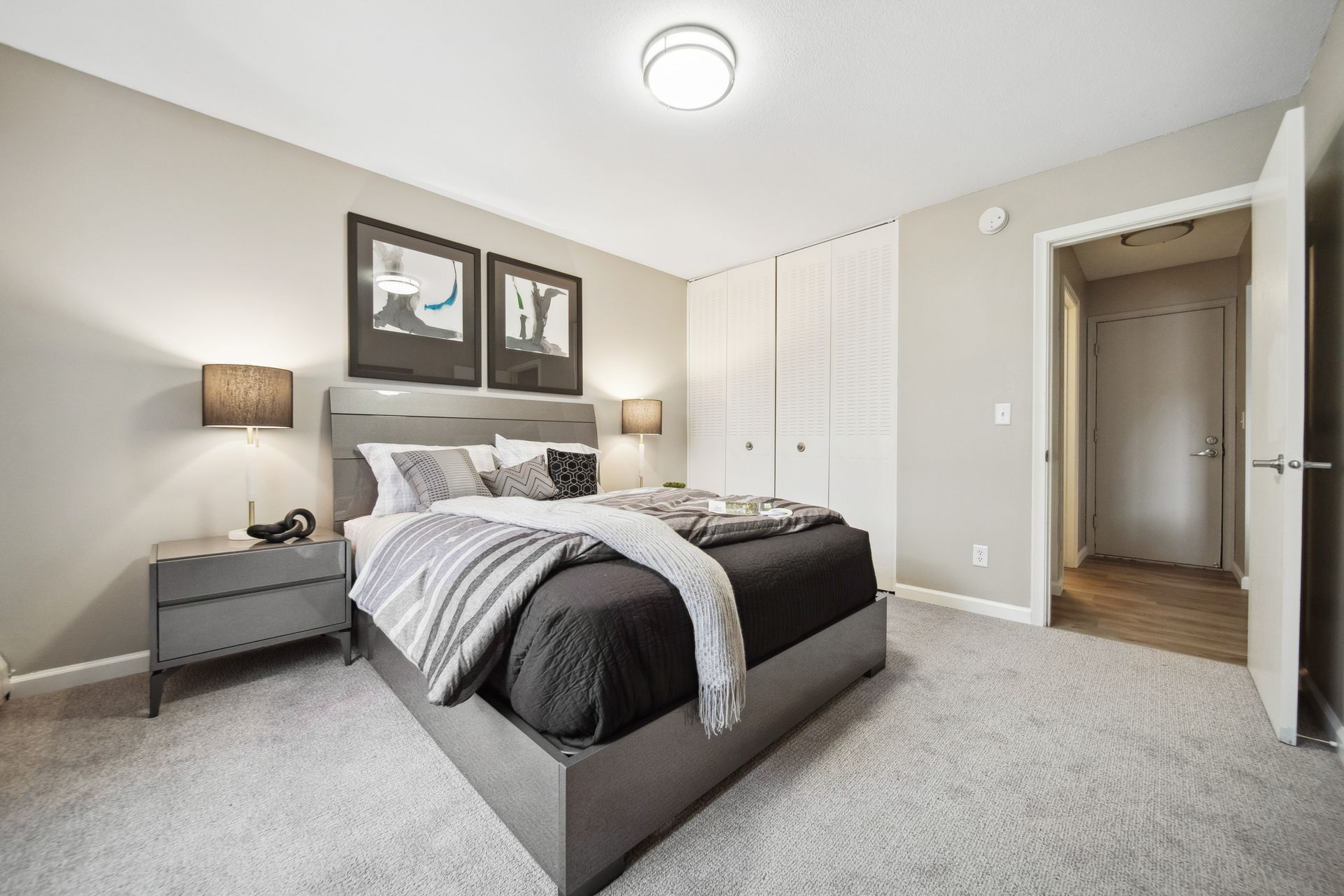 Modern bedroom with a gray upholstered bed, bedside lamps, wall art, and an open doorway to a hall.