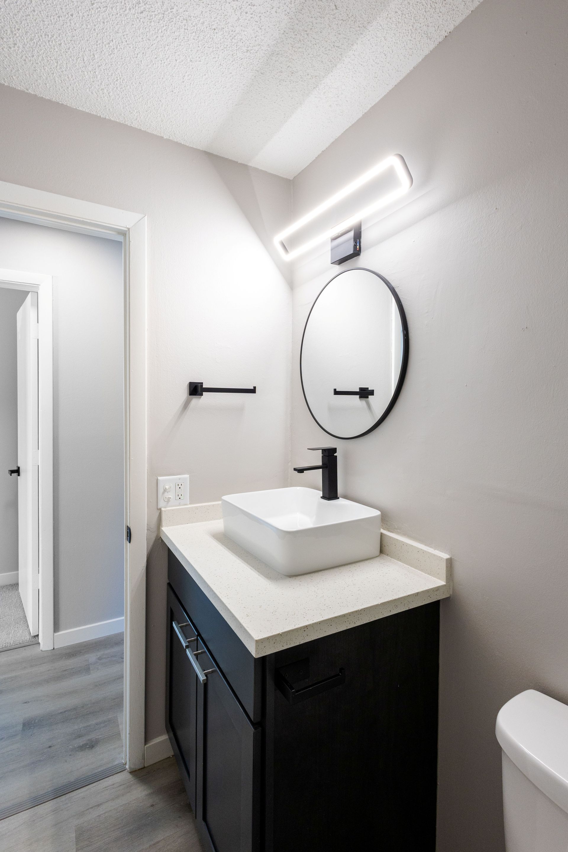Small bathroom vanity with round mirror, white sink, black cabinet, and open doorway to tiled hall.