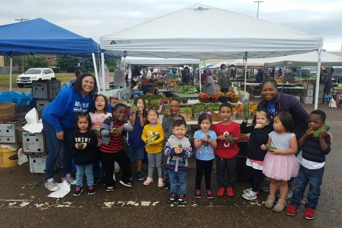 Group of children posing at an outdoor event under blue and white canopies, holding red cups.