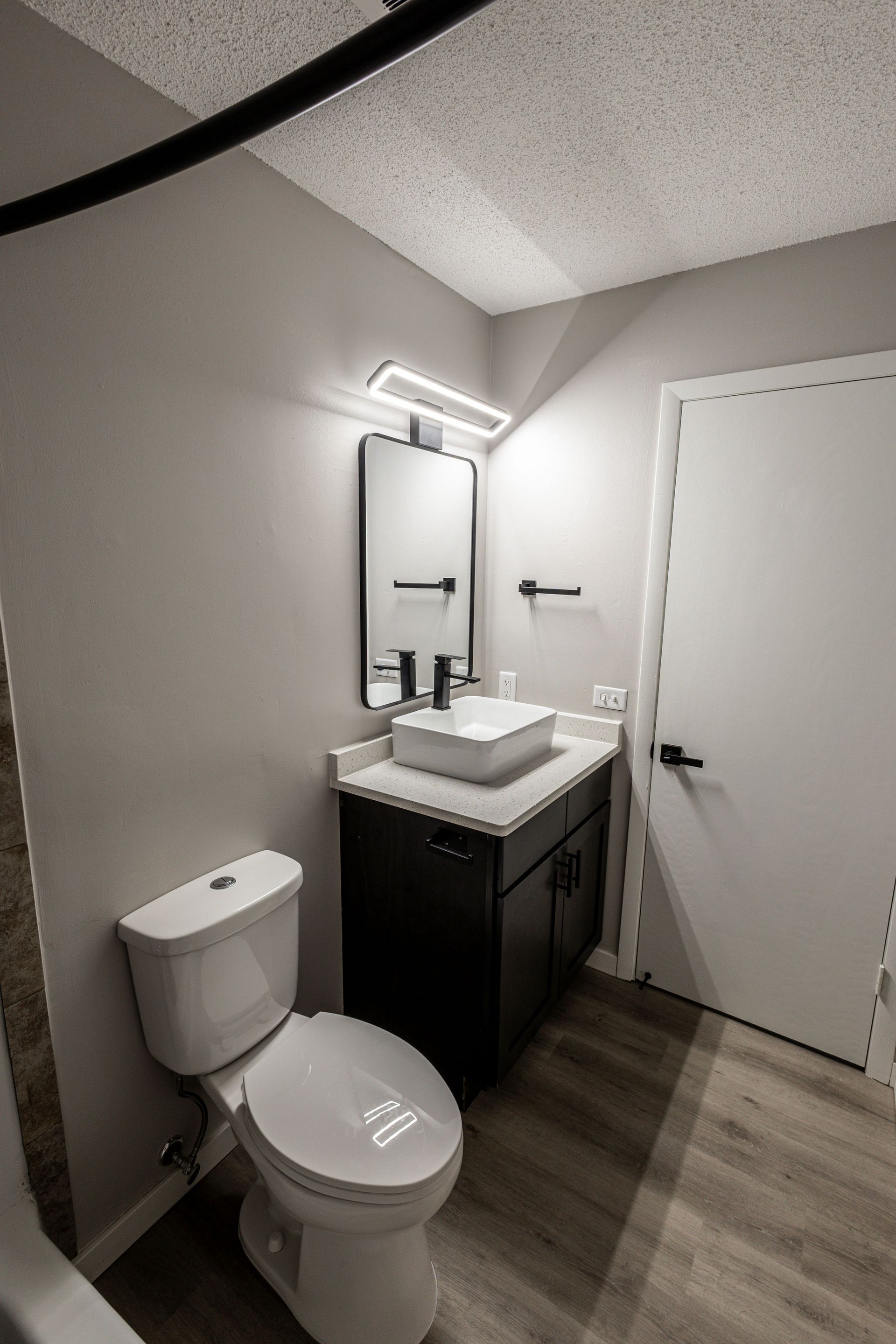 Small bathroom with toilet, sink vanity, mirror light, and white door on gray walls.