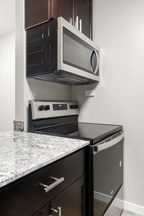 Compact kitchen corner with dark cabinets, granite countertop, microwave above stove, and refrigerator.
