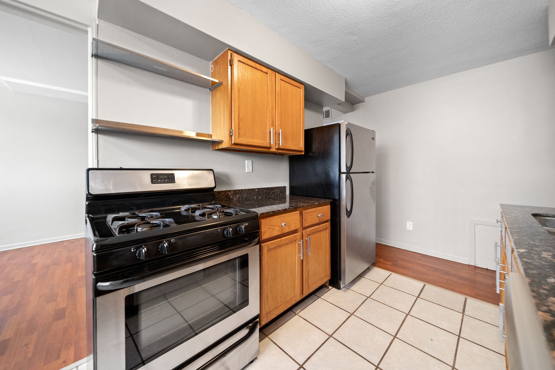 Compact kitchen with black stove, wood cabinets, stainless fridge, and tiled floor.