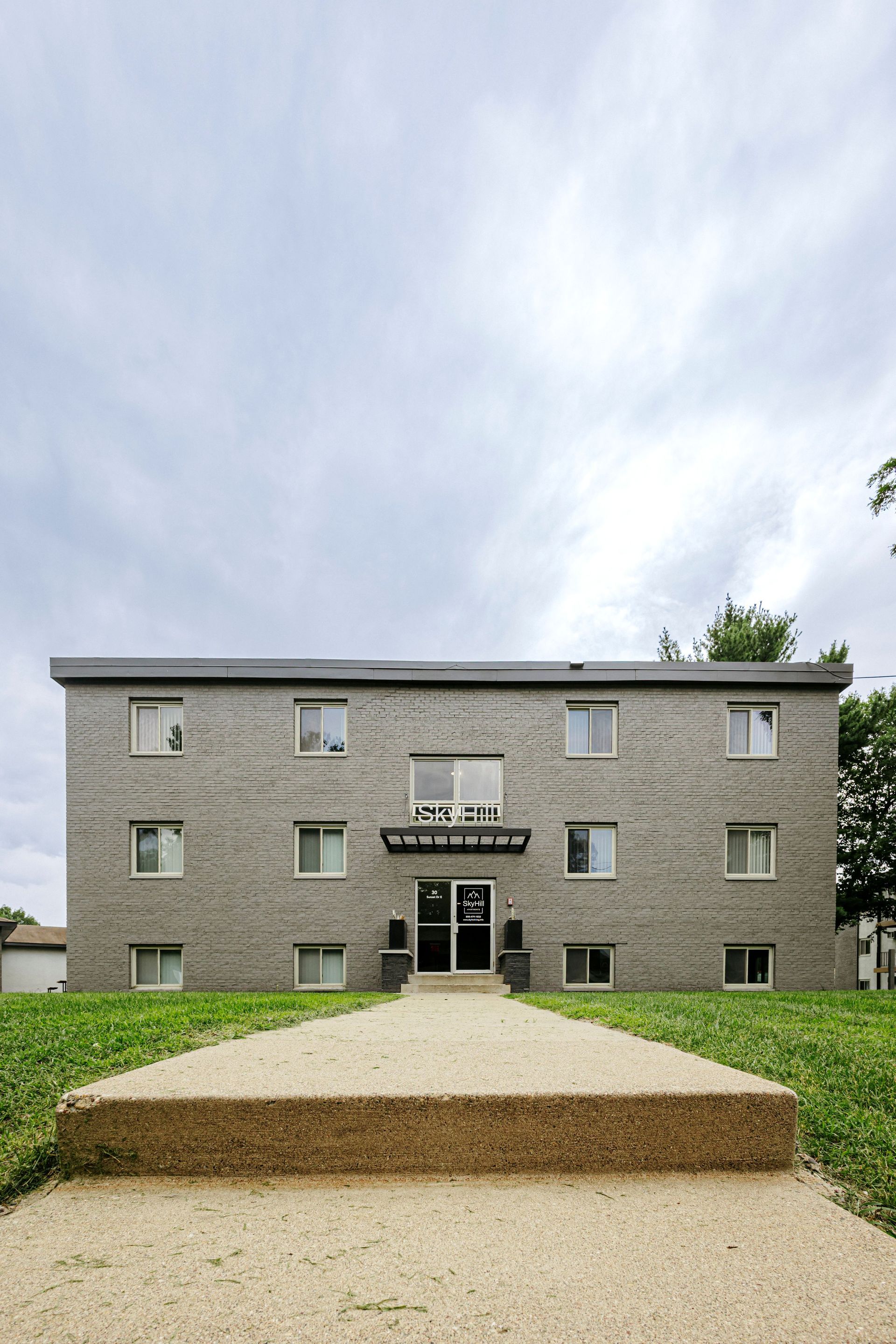 Gray brick apartment building with a central entrance and a concrete path under a cloudy sky