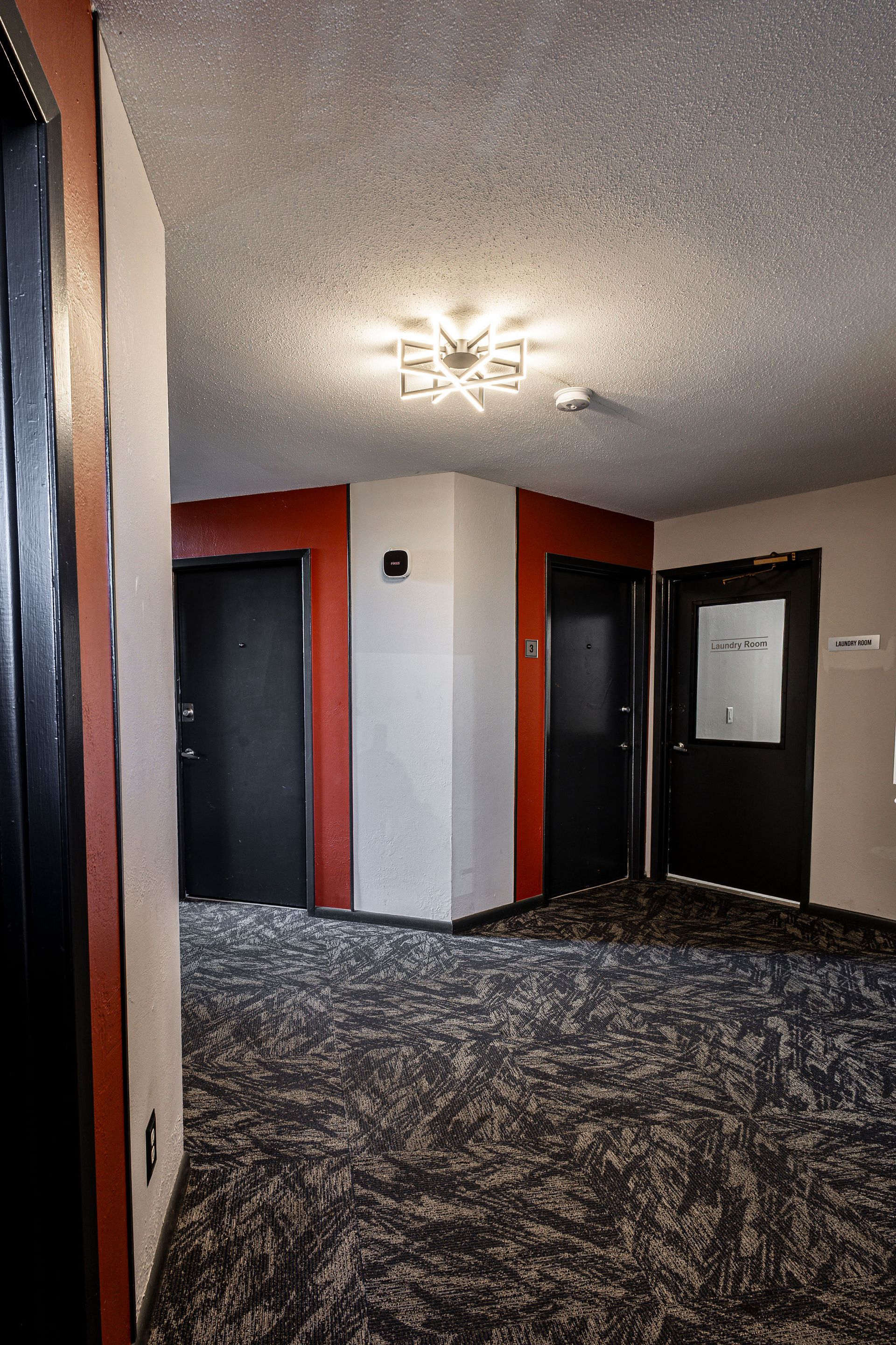 Dim apartment hallway with patterned carpet, red and beige walls, black doors, and a ceiling light.