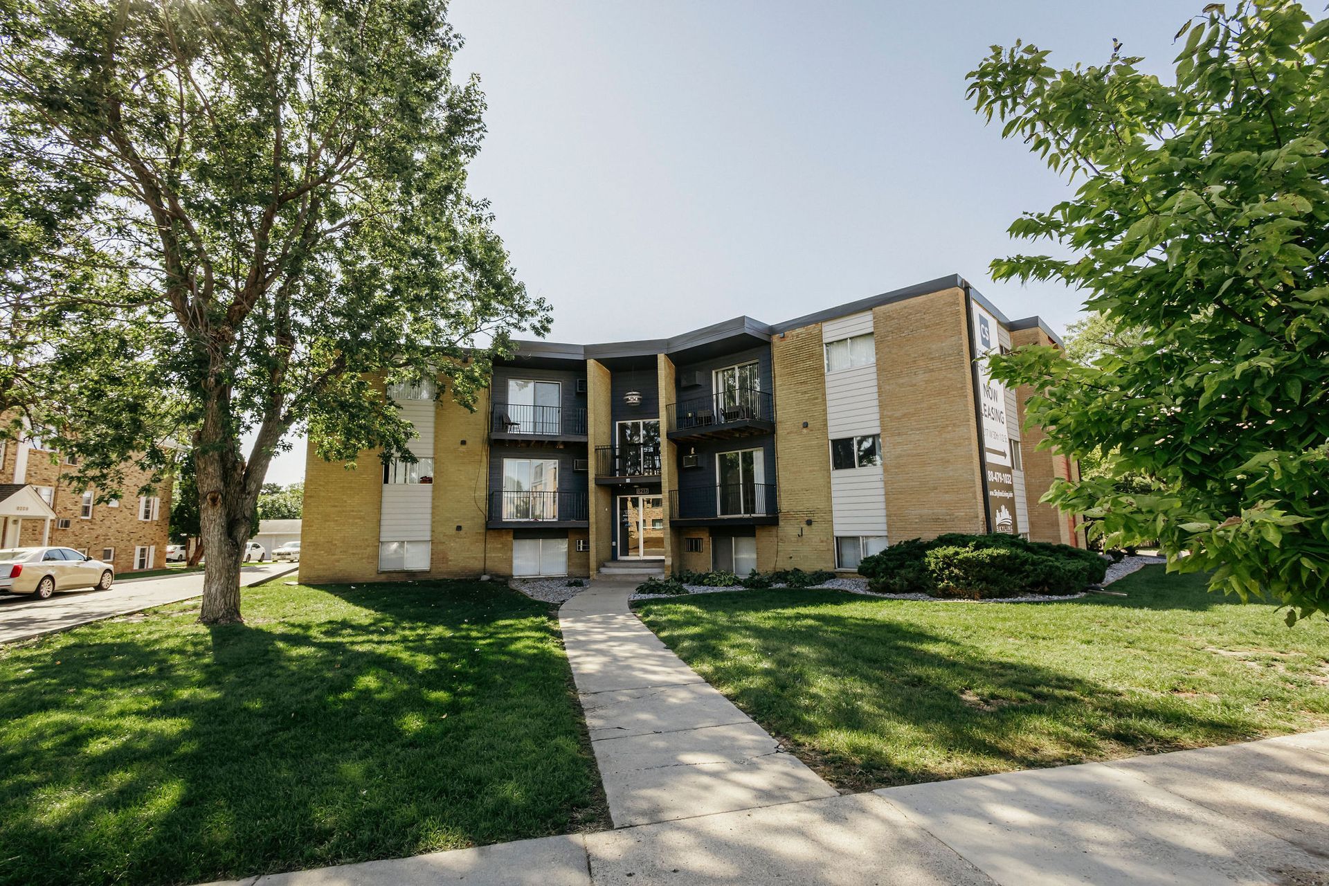 Apartment building with a central walkway, green lawn, and trees in front on a sunny day