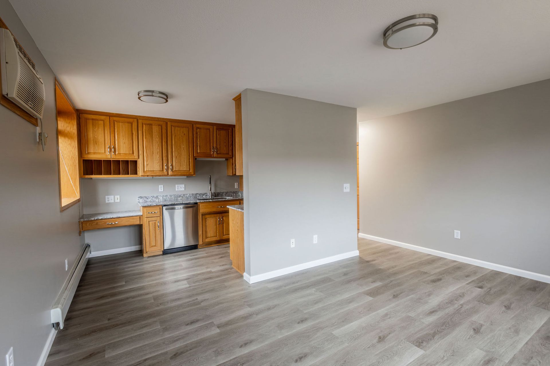 Empty apartment living area with kitchen, wood cabinets, gray walls, and light wood flooring