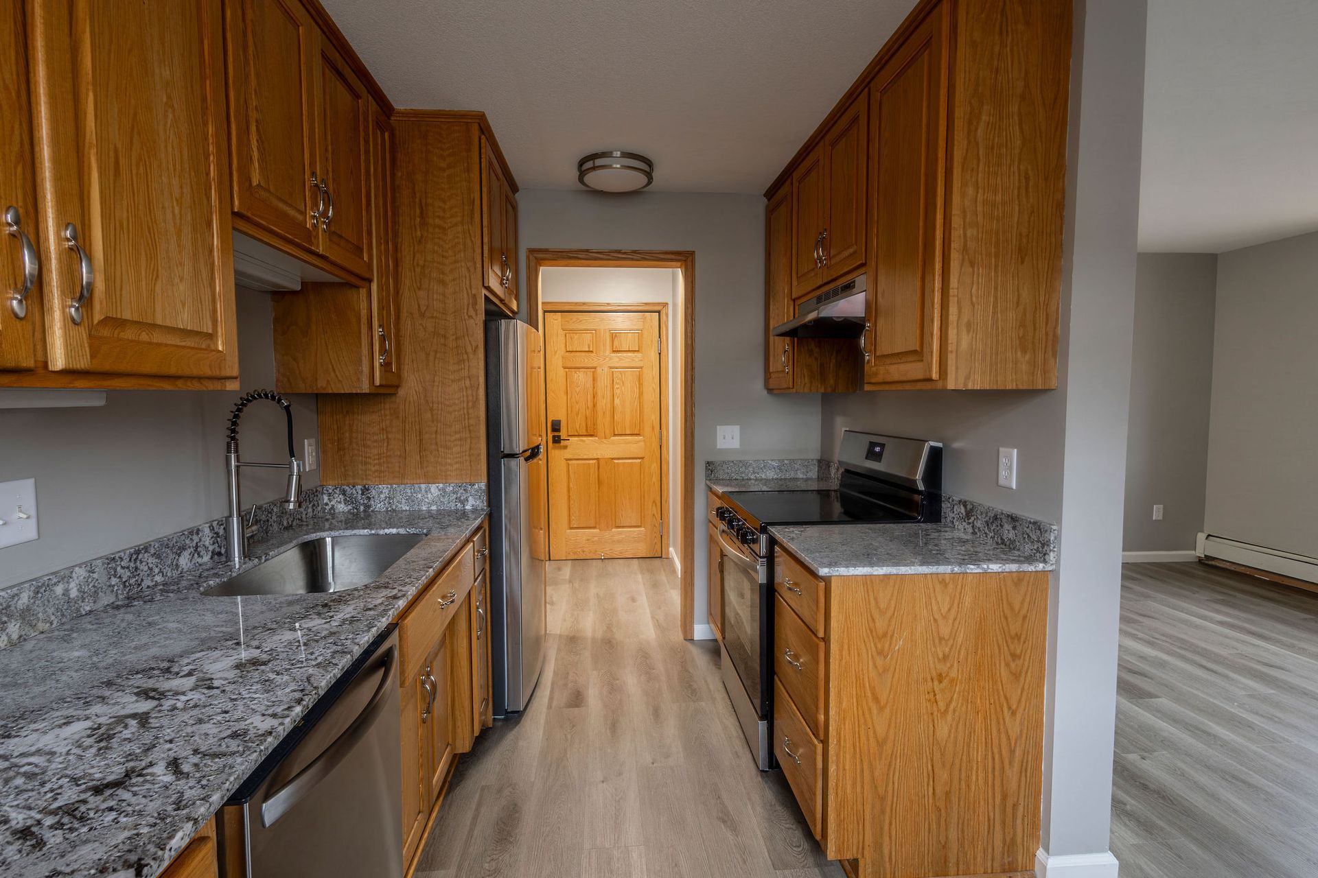 Narrow kitchen with wooden cabinets, granite countertops, stainless sink, and light wood flooring, looking toward a door.