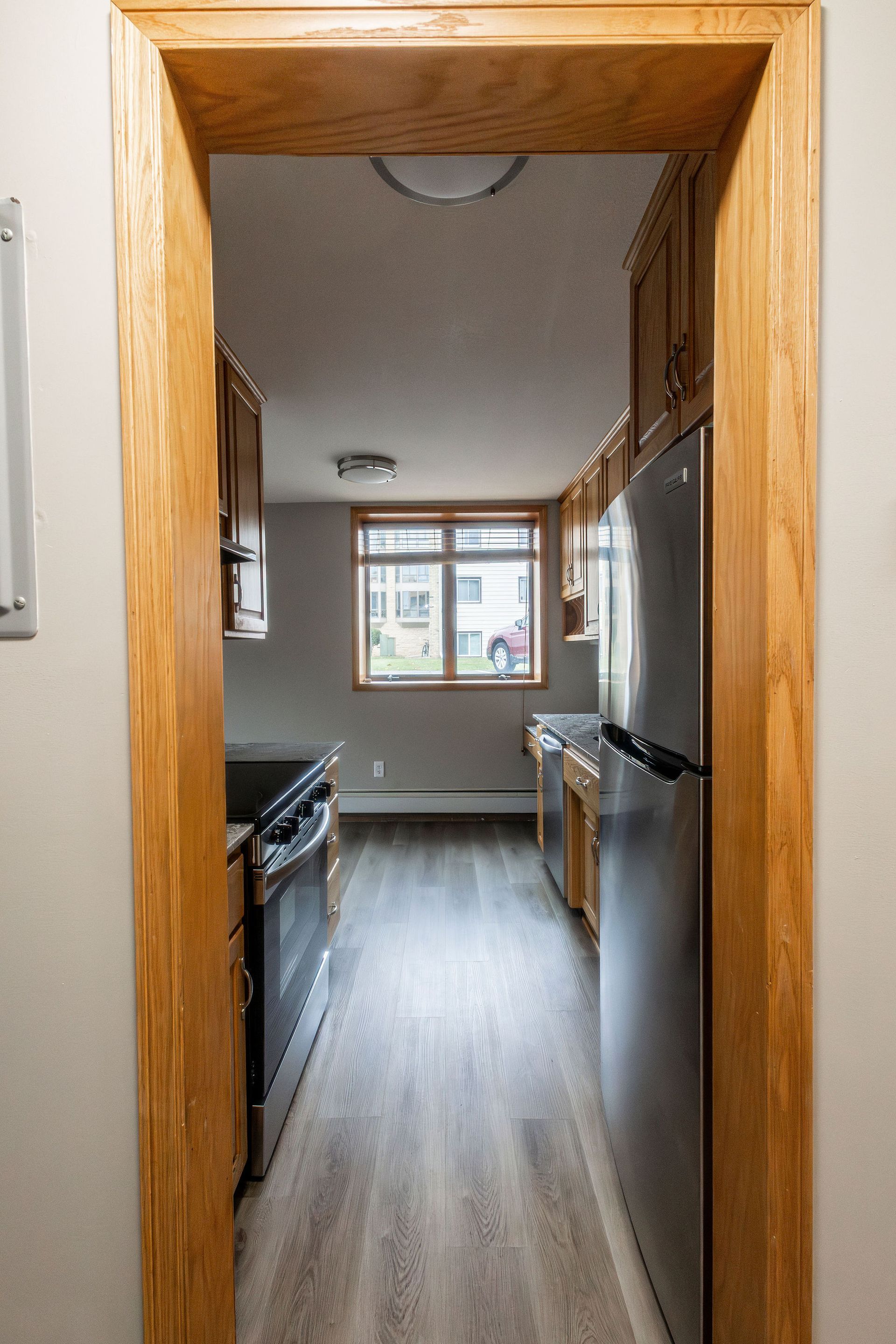 View through a wooden doorway into a narrow kitchen with wood floors, white cabinets, and a window at the far end