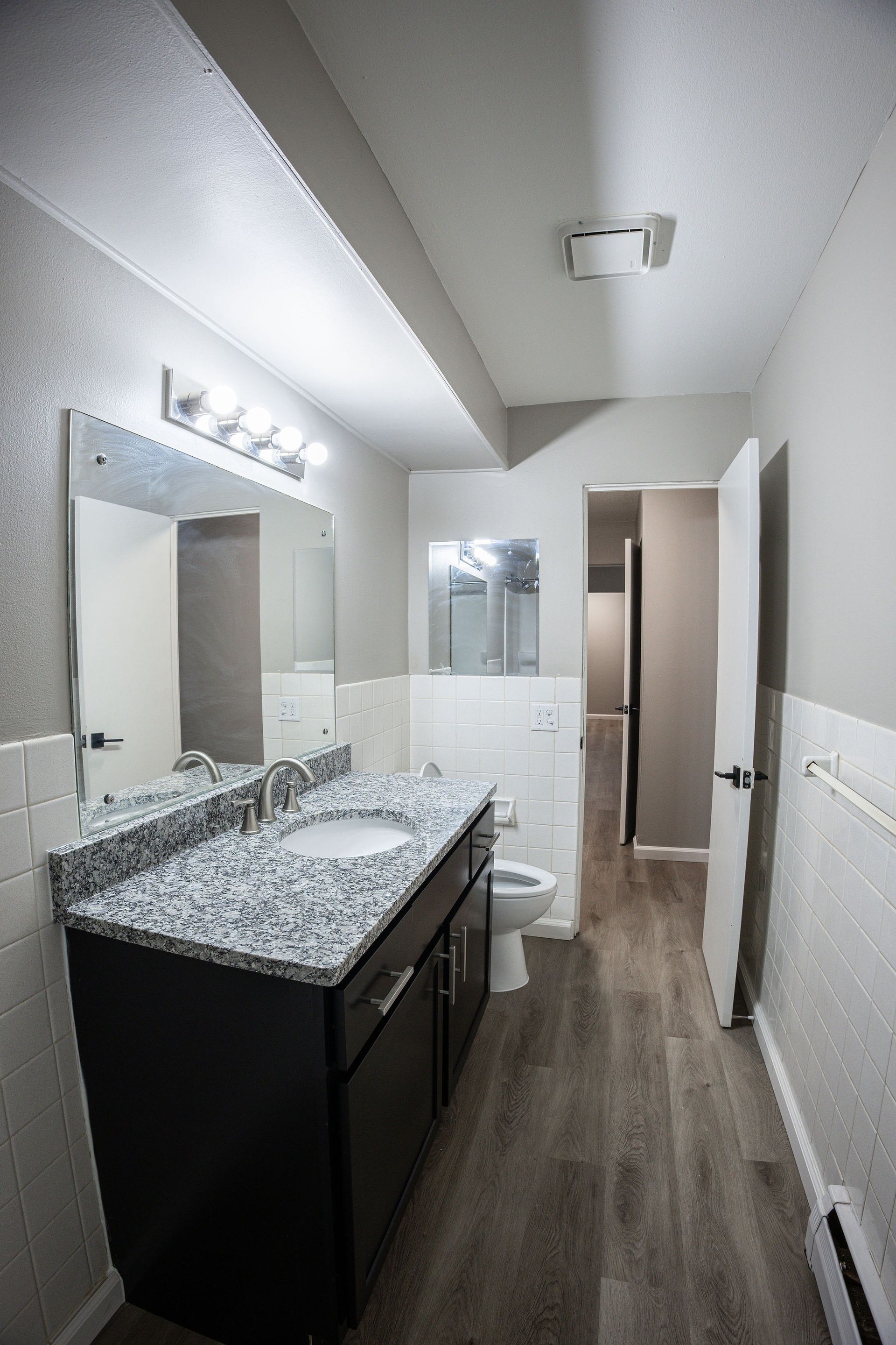 Small bathroom with granite vanity, large mirror, toilet, and walk-in shower in a narrow hallway.
