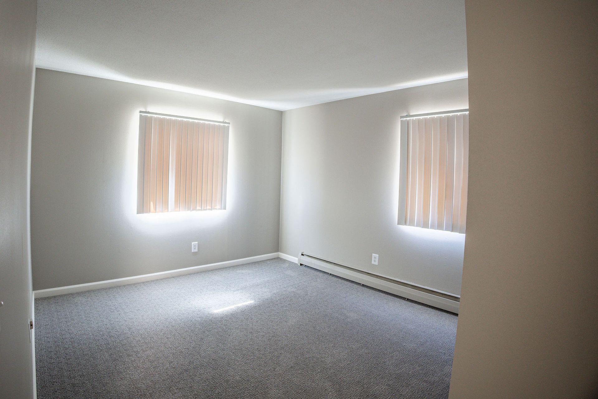 Empty gray-carpeted room with beige walls and two windows with blinds