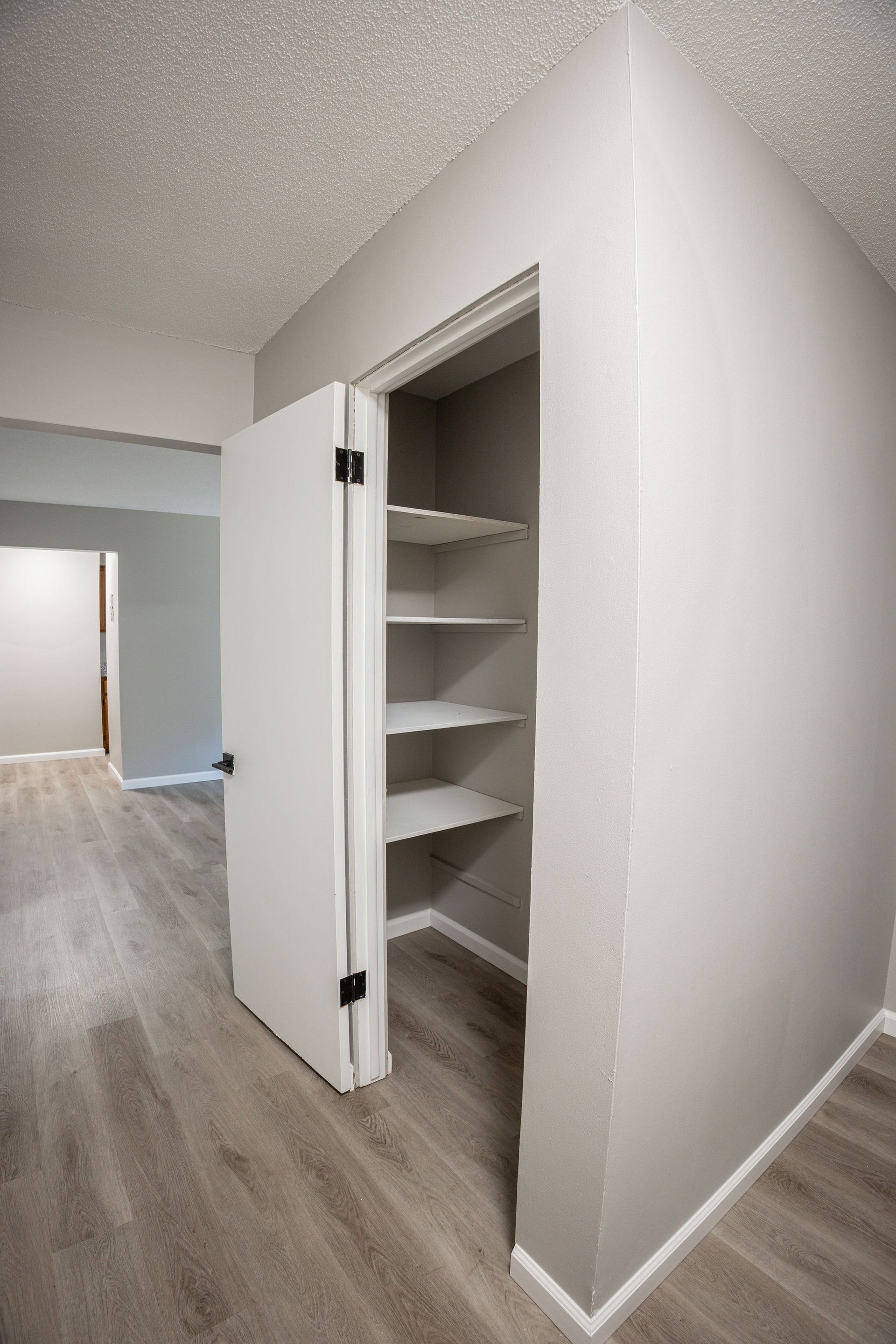 Open pantry closet with white shelves in a hallway, beside a room with wood-look flooring.