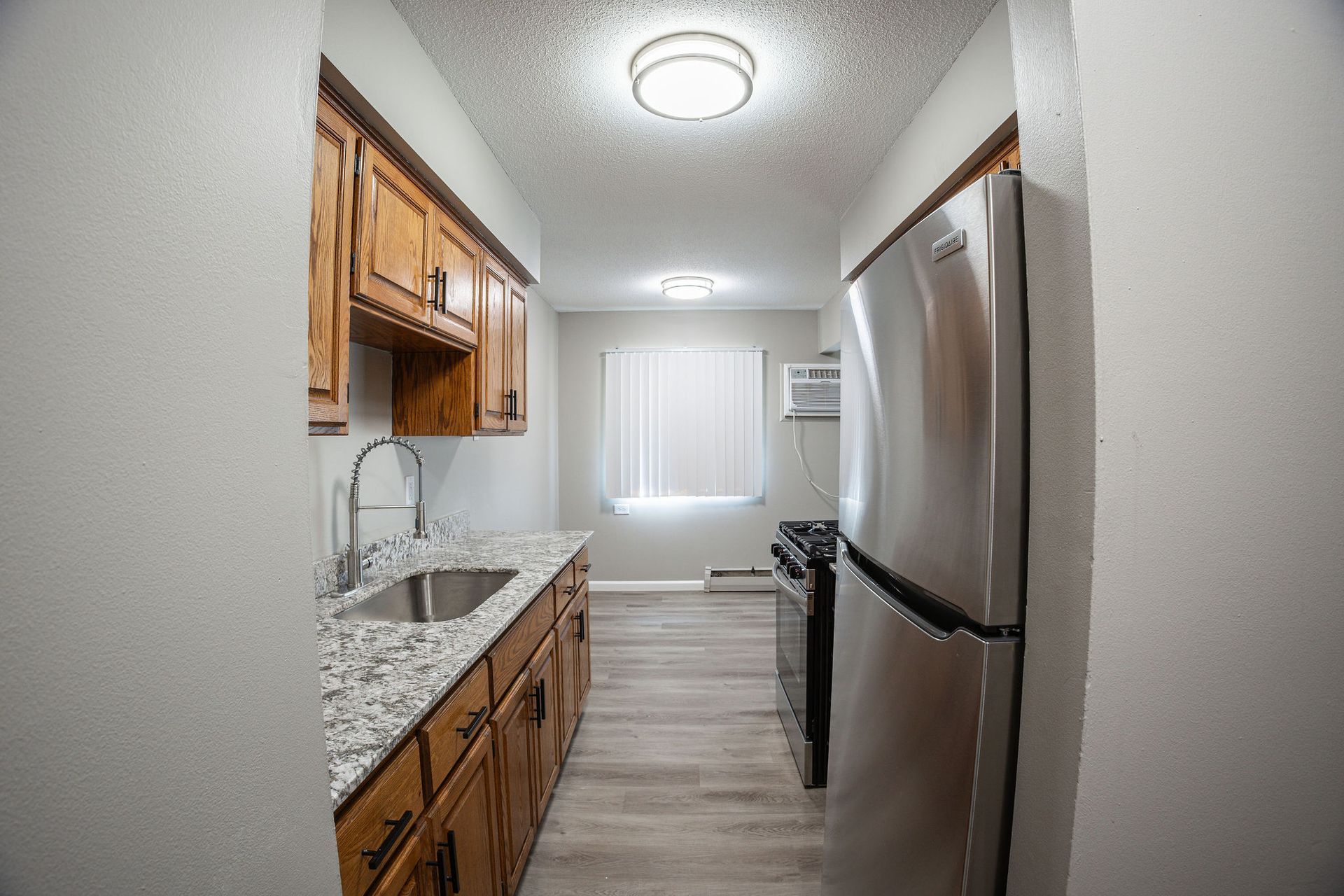 Narrow galley kitchen with wood cabinets, granite counters, stainless fridge, and window at the end.