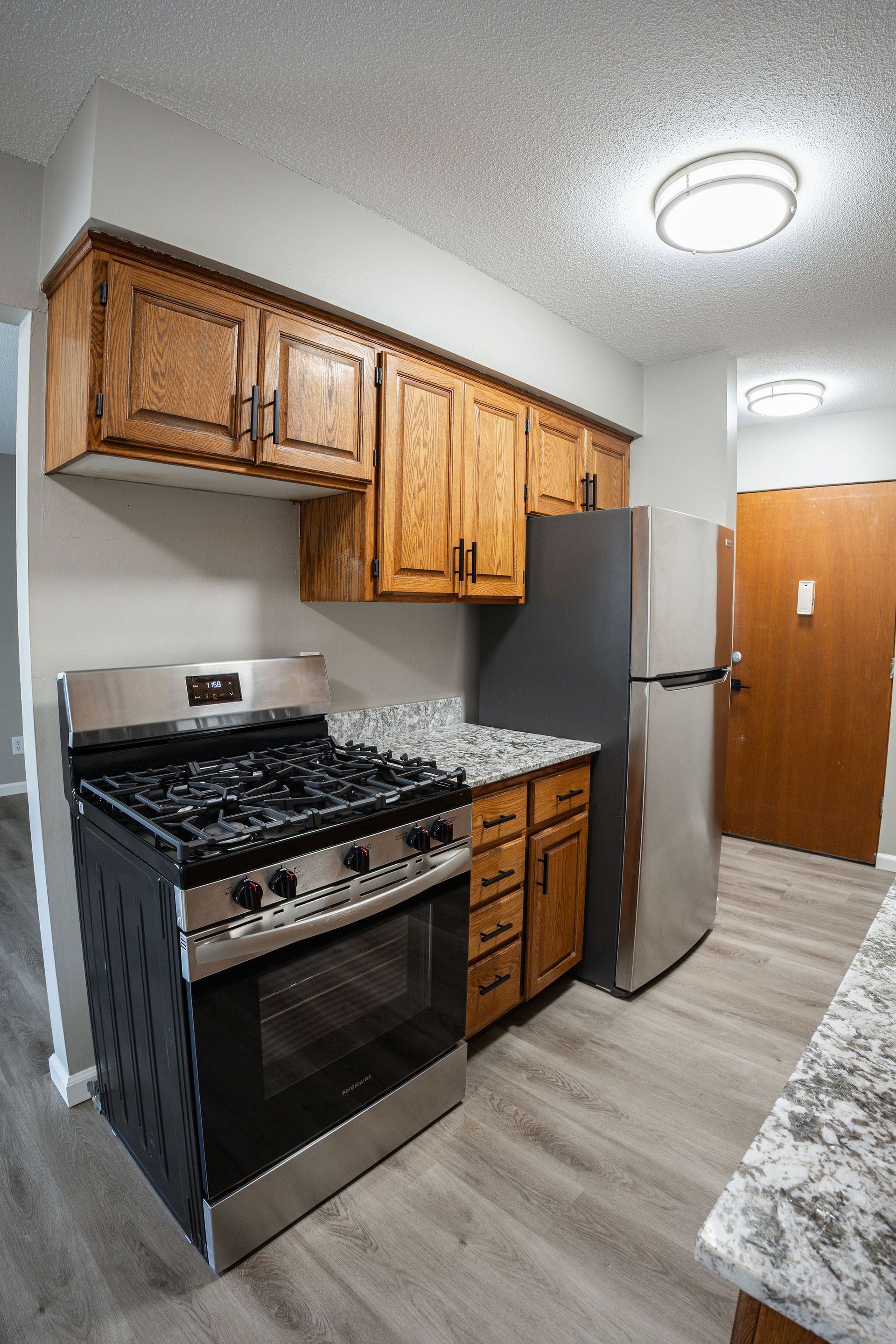 Small kitchen with wood cabinets, gas stove, gray fridge, and light tile floor.