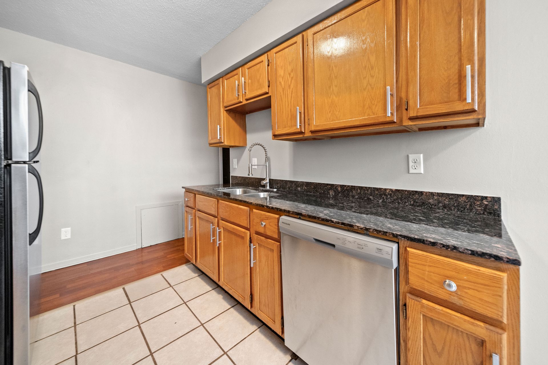 Empty kitchen with wooden cabinets, black countertop, stainless dishwasher, and beige tile floor