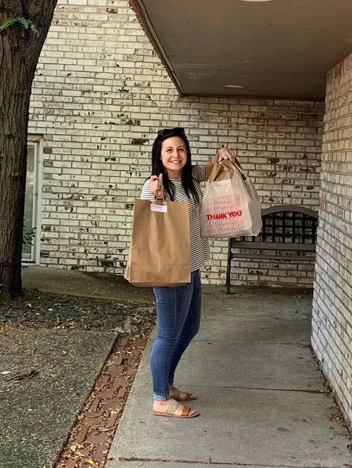 Person standing on a covered walkway, smiling and holding two shopping bags in front of a white brick wall.