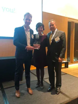 Three people in formal wear holding an award on a conference stage with a projection screen behind them