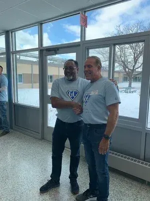 Two men in gray T-shirts shake hands in a bright indoor lobby with large windows.