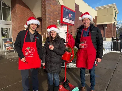 Three people in Santa hats and red aprons stand outside a store, smiling and holding donation buckets.