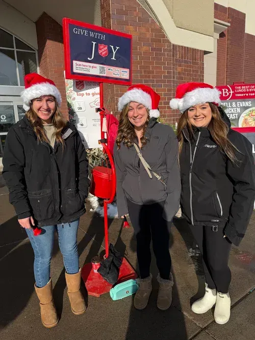 Three people in Santa hats standing by a red donation stand outside a brick store