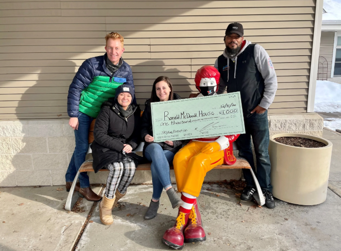 Four people posing outdoors with a large check, seated by a house wall on a sunny day