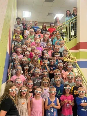 Group of children on school stairs wearing colorful sunglasses, with two adults at the top.