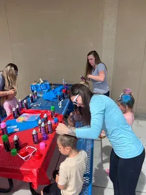 People gathered around a red tablecloth at a craft station, arranging colorful supplies and bottles.