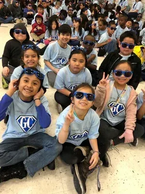 Smiling children seated outdoors, many wearing sunglasses and matching shirts, posing for a group photo