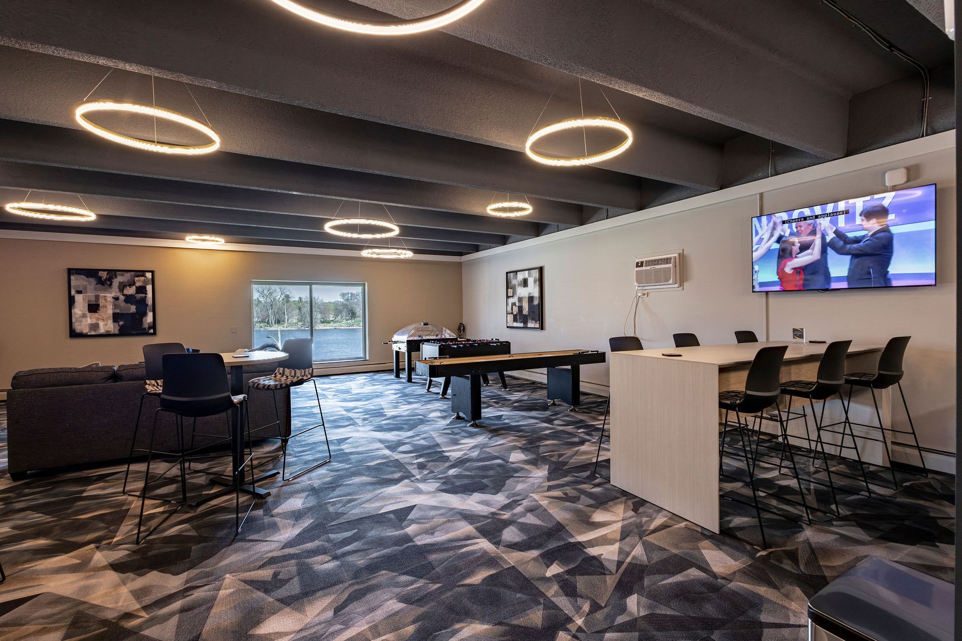 Modern lounge with patterned carpet, circular ceiling lights, chairs, tables, and wall-mounted TVs.