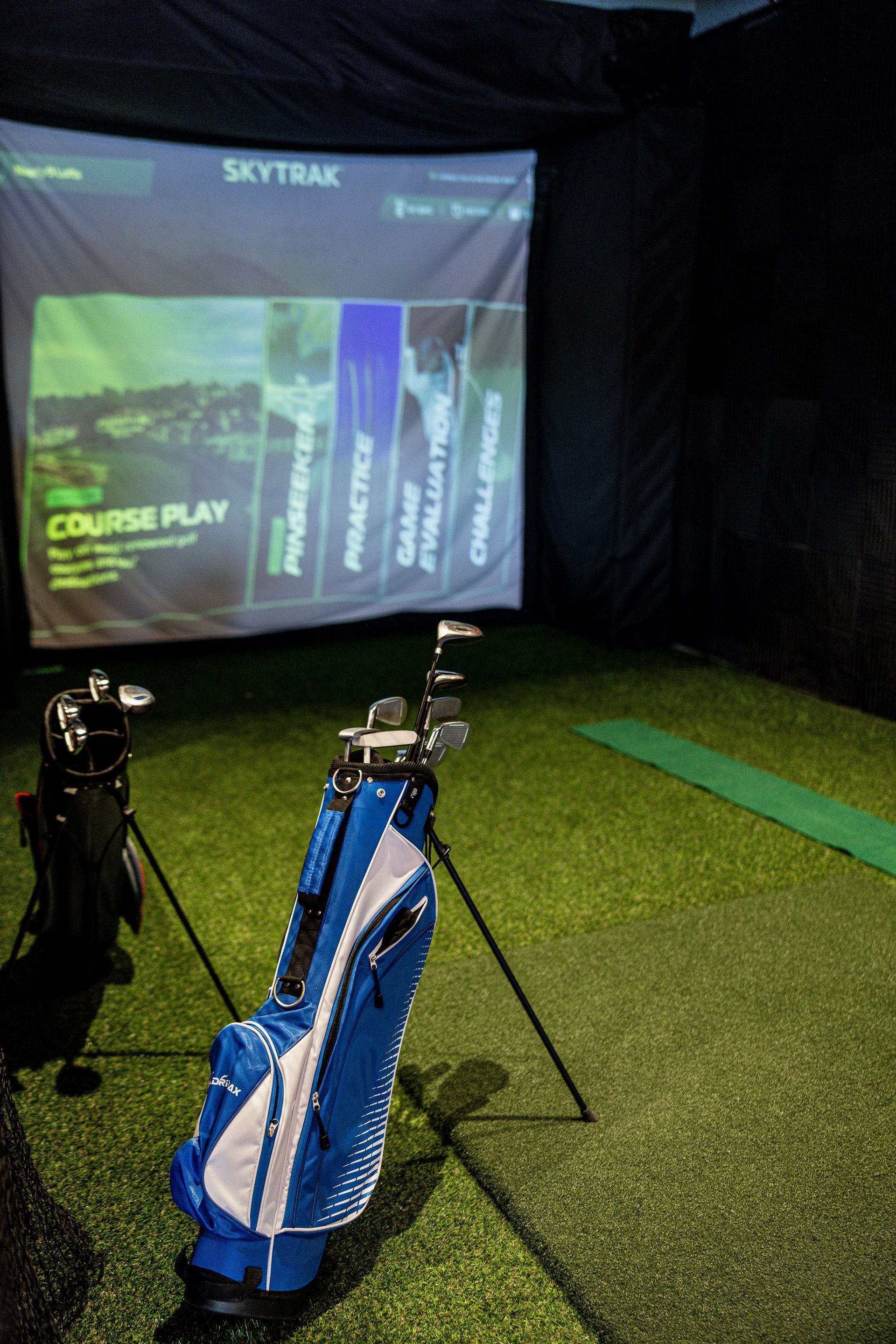 Indoor golf simulator bay with two golf bags on turf and a projected course on the screen.