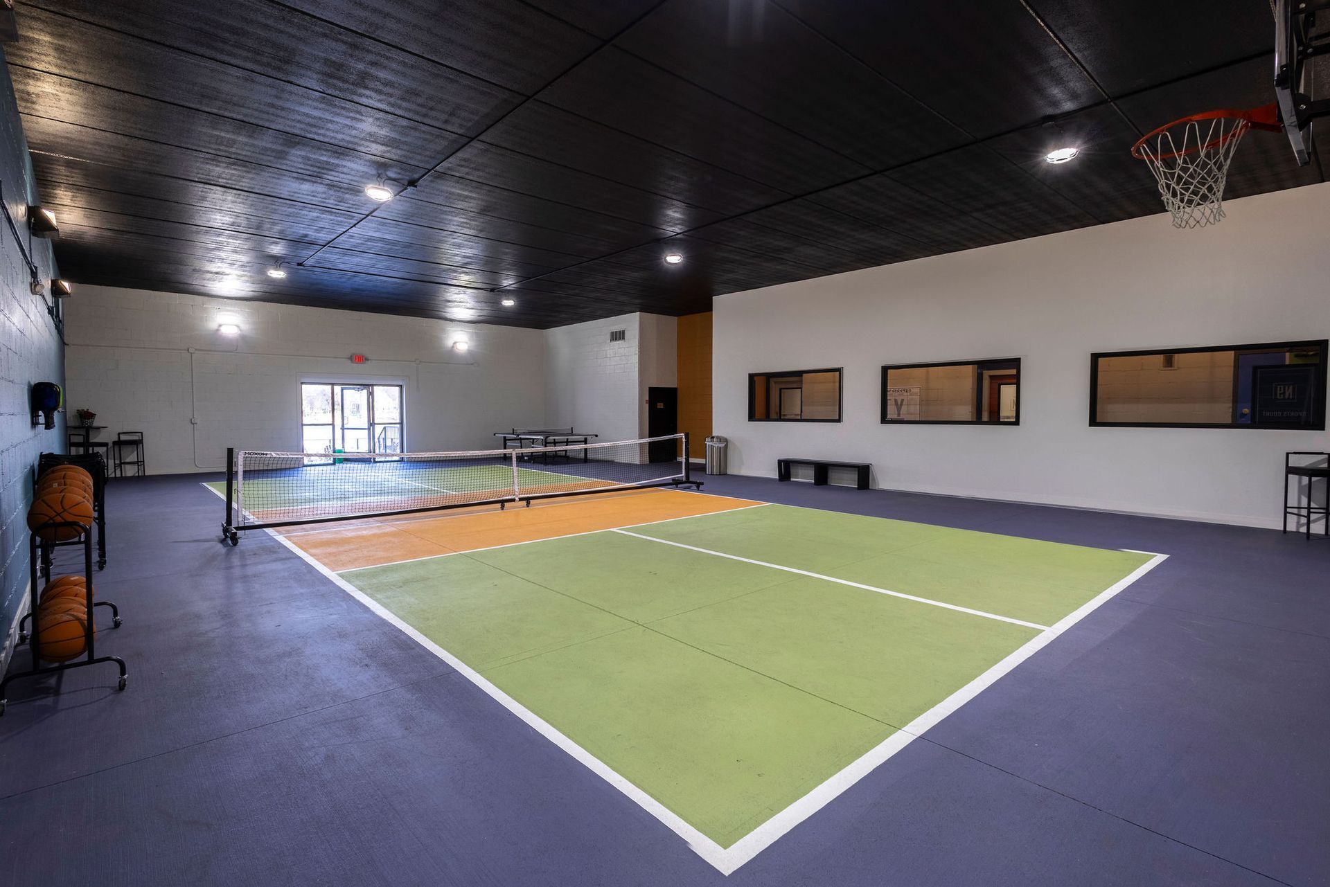 Indoor sports court with green playing surface, orange center lane, and basketball hoop in a bright gymnasium