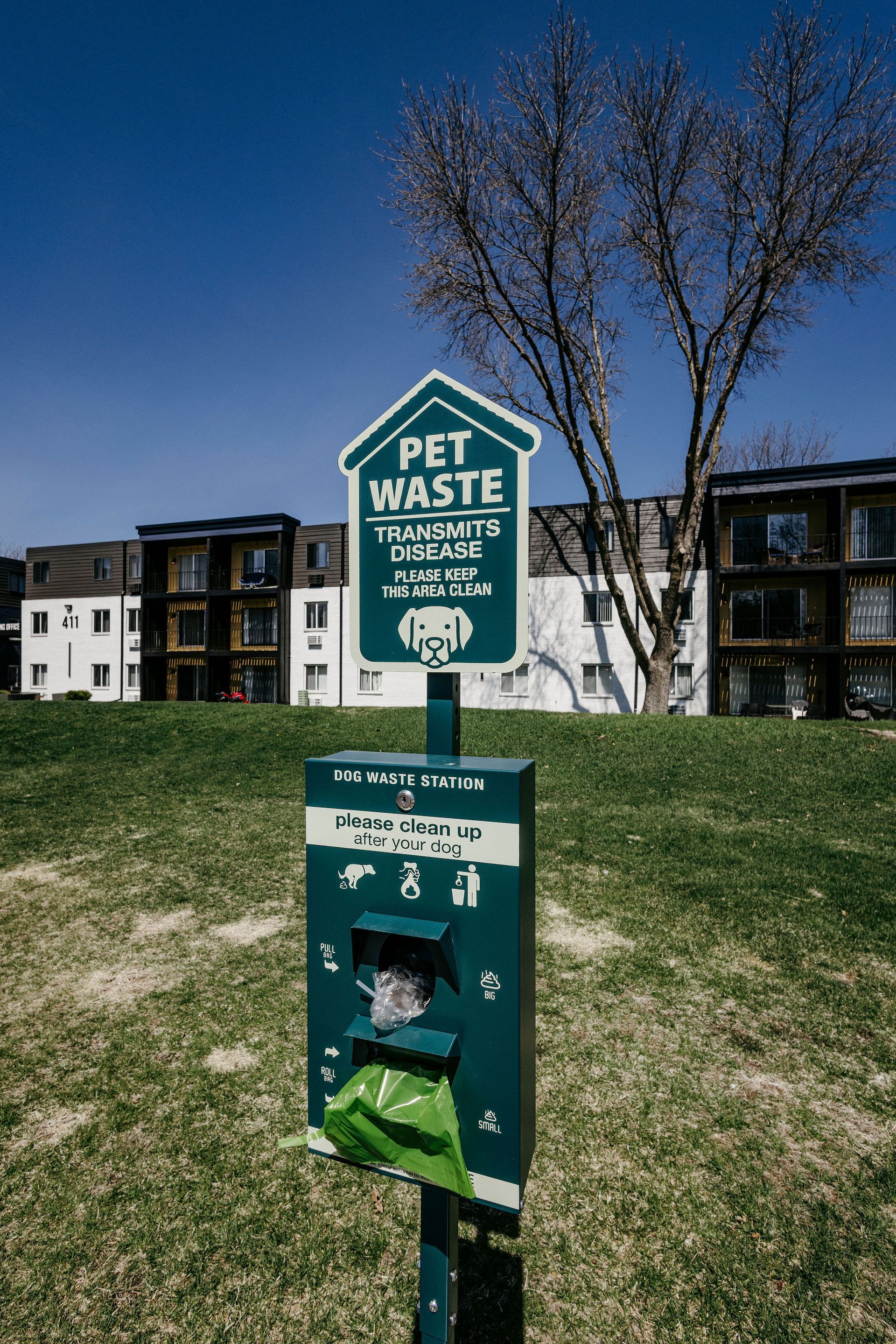 Green pet waste station in a grassy area near apartment buildings under a blue sky.