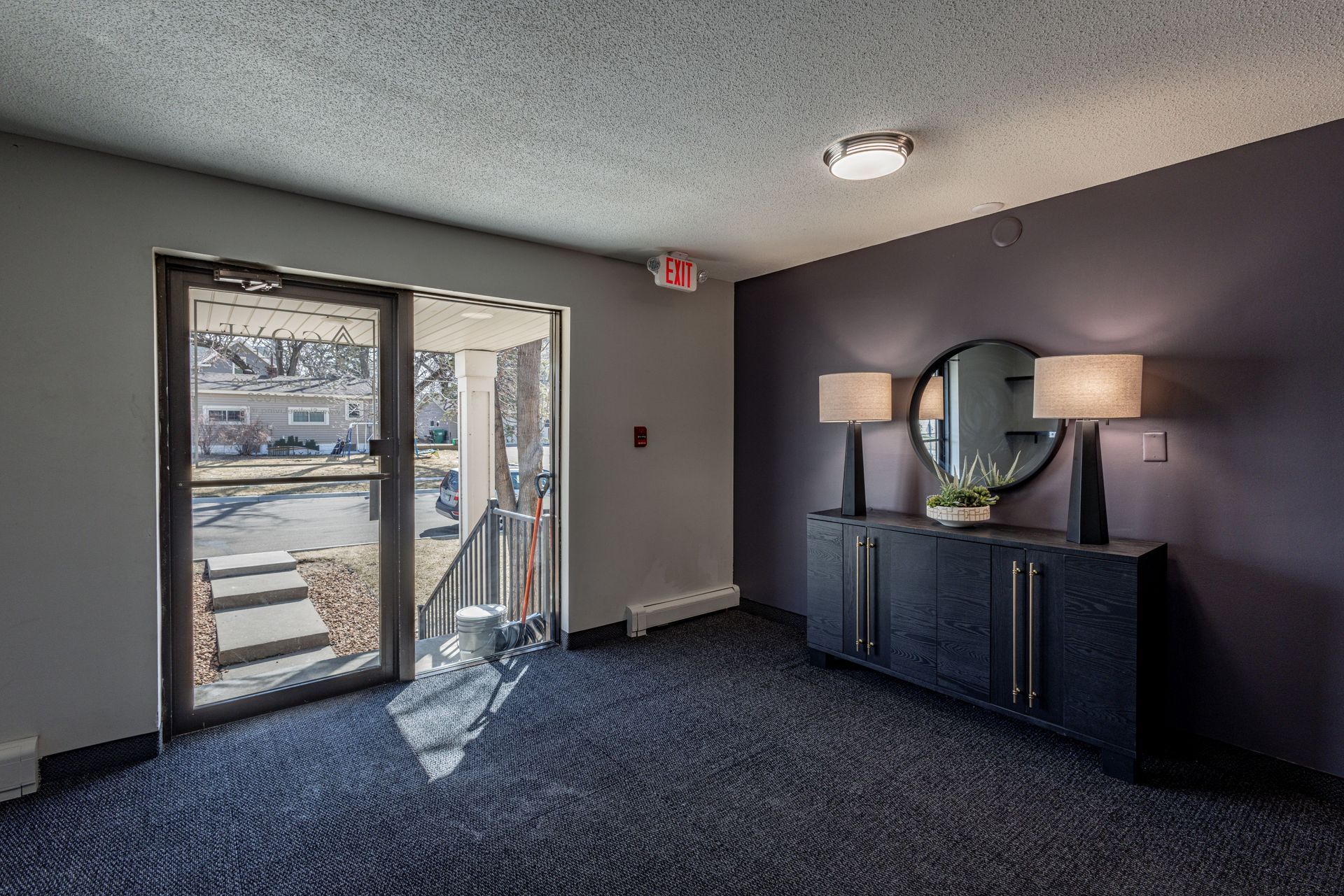 Empty living room with gray carpet, dark accent wall, sliding glass door, and a console with lamps and mirror.