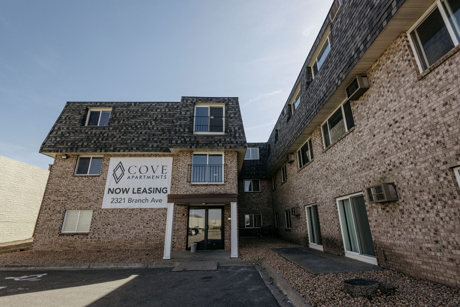 Stone apartment buildings with a “now leasing” sign above the entrance and parking lot in front