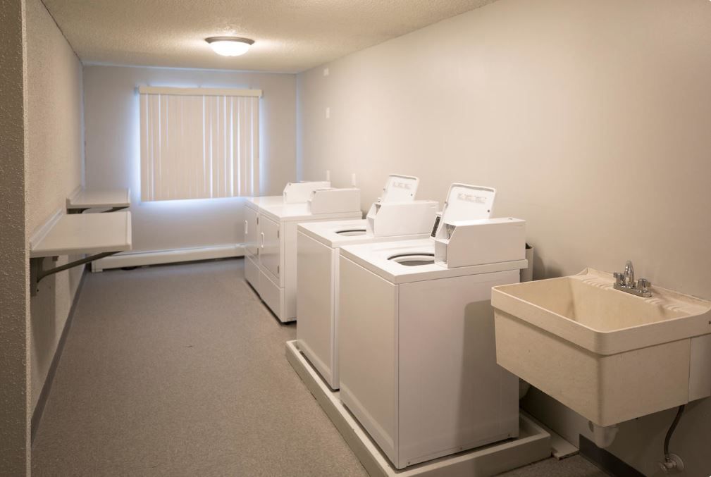 Laundry room with rows of white washing machines and a utility sink under a window.