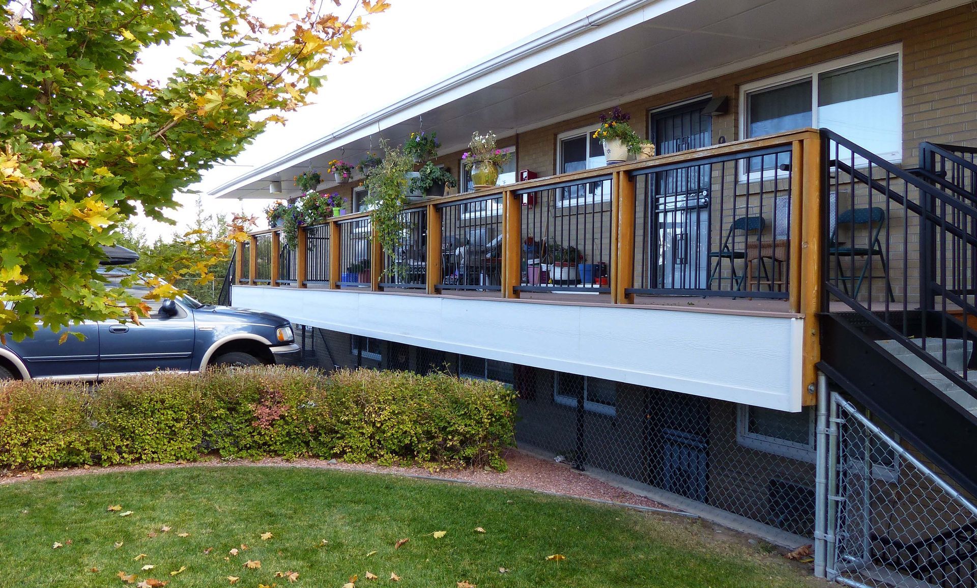 Apartment building with a long balcony, black railings, and a parked car beside a lawn and shrubs