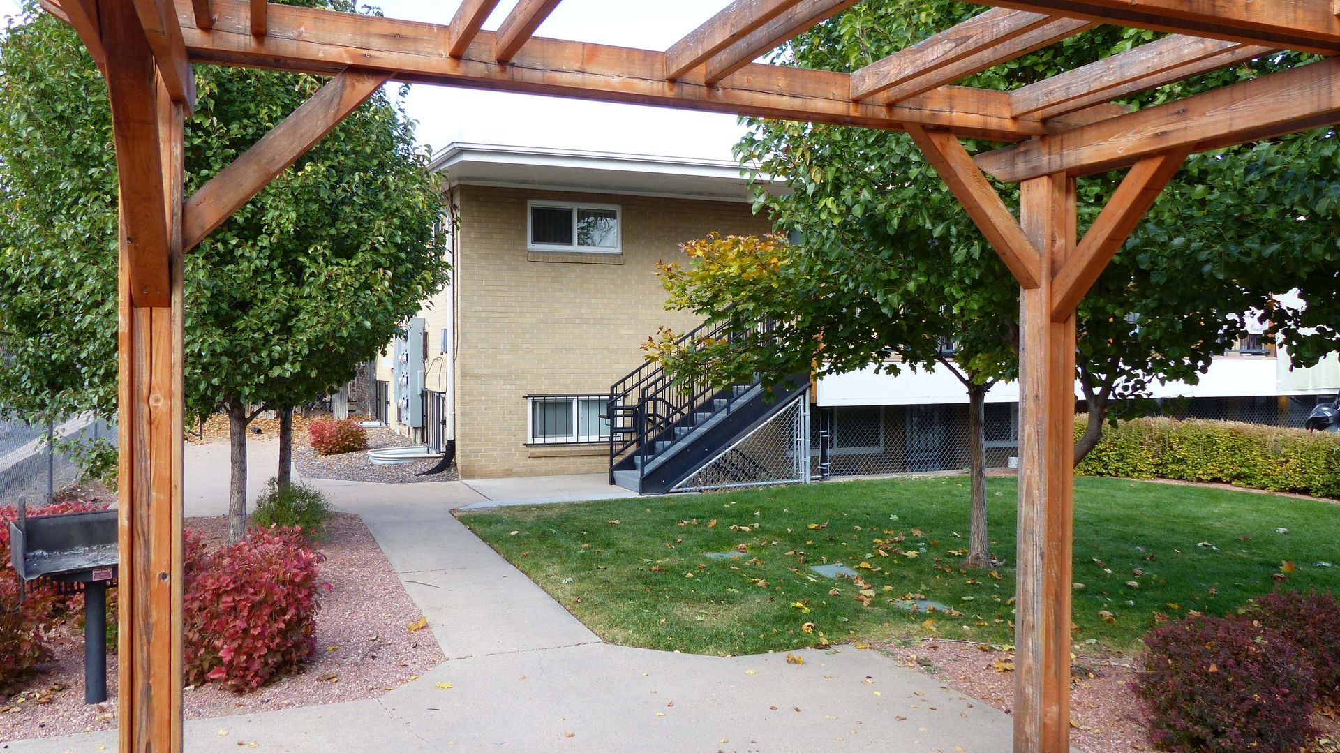 Wooden pergola framing a walkway to a tan apartment building with stairs and green lawn